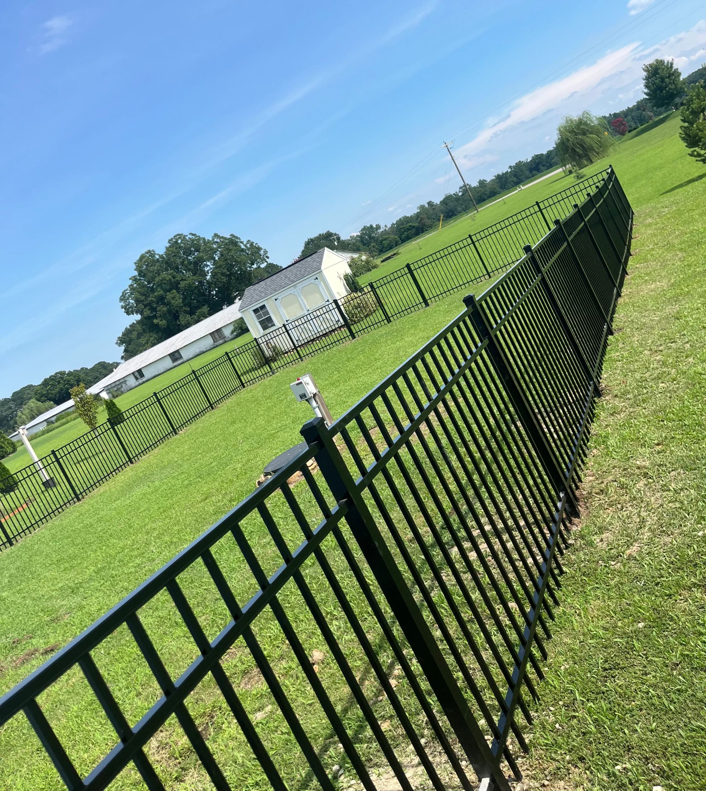 A long black aluminum picket fence stretching across a large green field toward a white shed under a clear blue sky