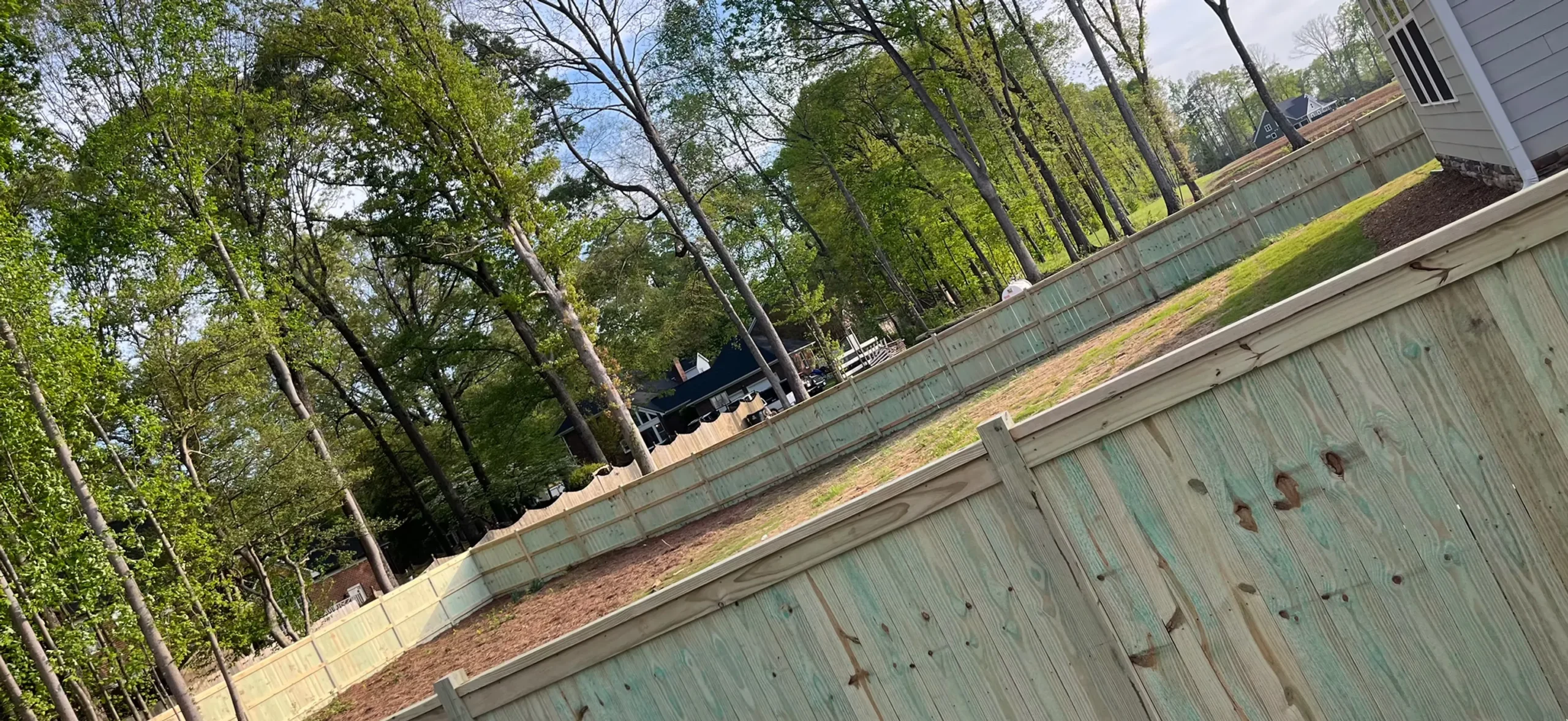 A long wooden fence with a light green tint encloses a large, grassy backyard surrounded by tall trees